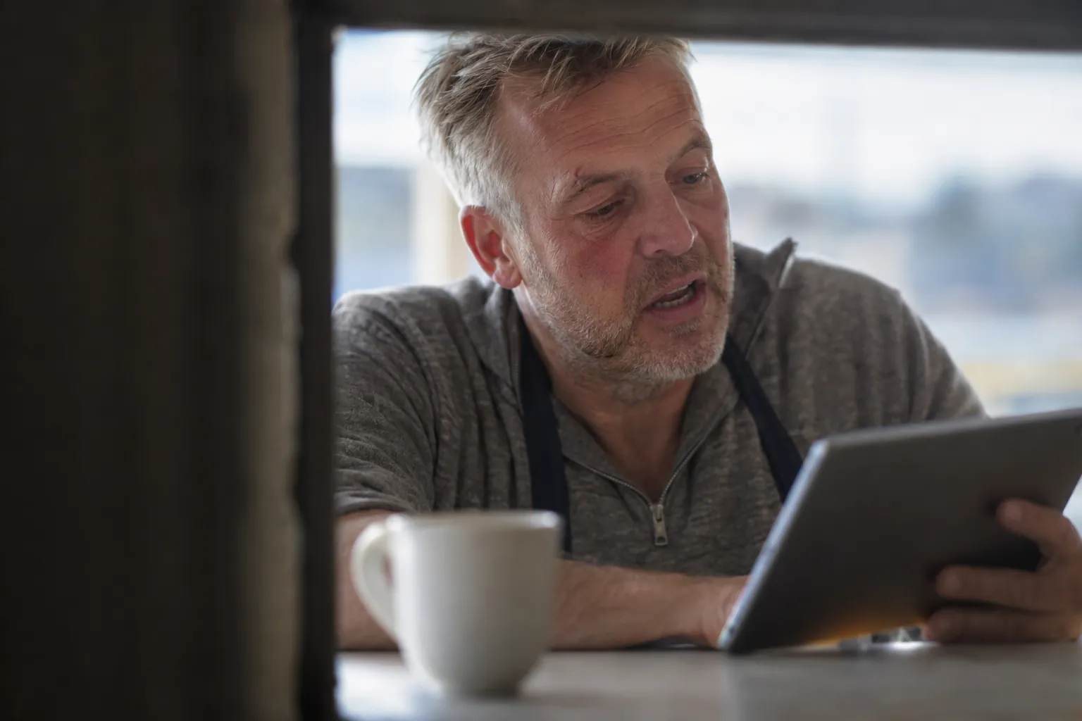 Host in an apron checking bookings on a tablet over coffee